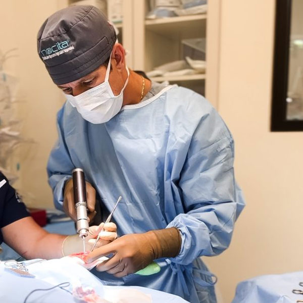 A surgeon wearing a grey cap, surgical mask, and blue scrubs performs a medical procedure using a handheld instrument. Another individual, partially visible, assists. Medical equipment and supplies are visible in the background.
